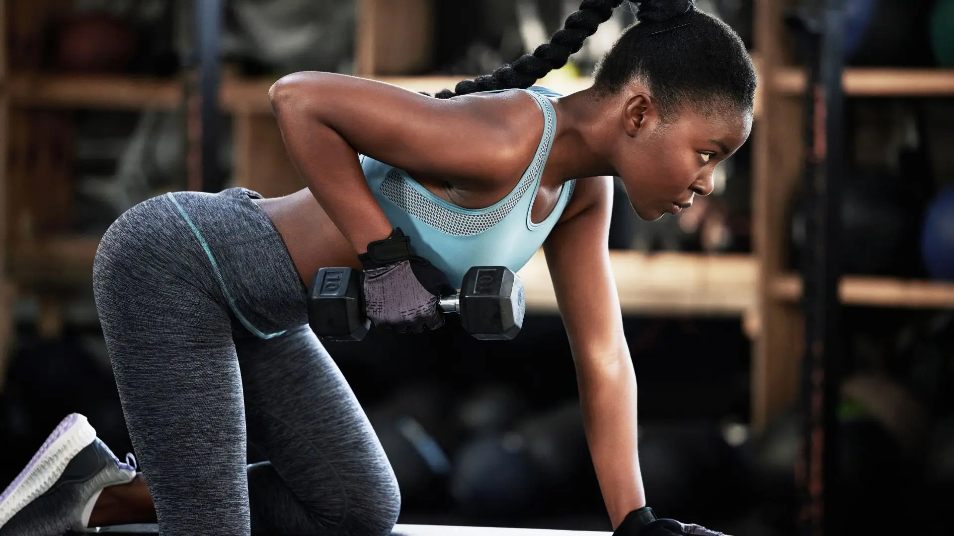 Athlete performing a bent-over dumbbell row in a gym, wearing a light blue sports bra and gray leggings.