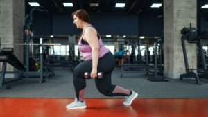 Should I do cardio or strength training for fat loss? - Falls Church Personal Training Woman in a pink athletic top and black leggings performs a lunge while holding dumbbells in a gym setting.