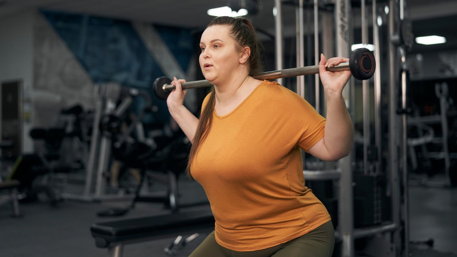 Woman in a pink athletic top and black leggings performs a lunge while holding dumbbells in a gym setting.