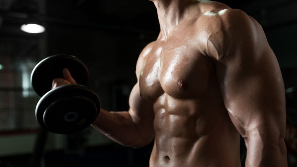 Muscular, shirtless man lifting a dumbbell in a dimly lit gym, focusing on upper body strength.