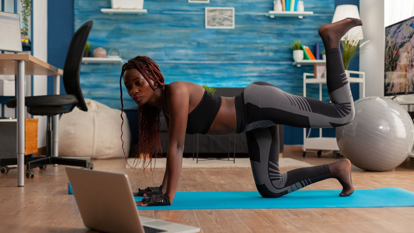 Woman in a black-and-gray active outfit performs a bird-dog exercise on a blue mat, following a workout video on a laptop.